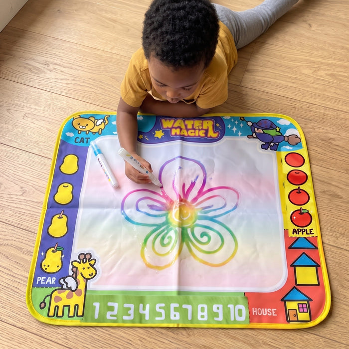 A child lies on the floor drawing a big rainbow-colored flower on the mat with a water pen.