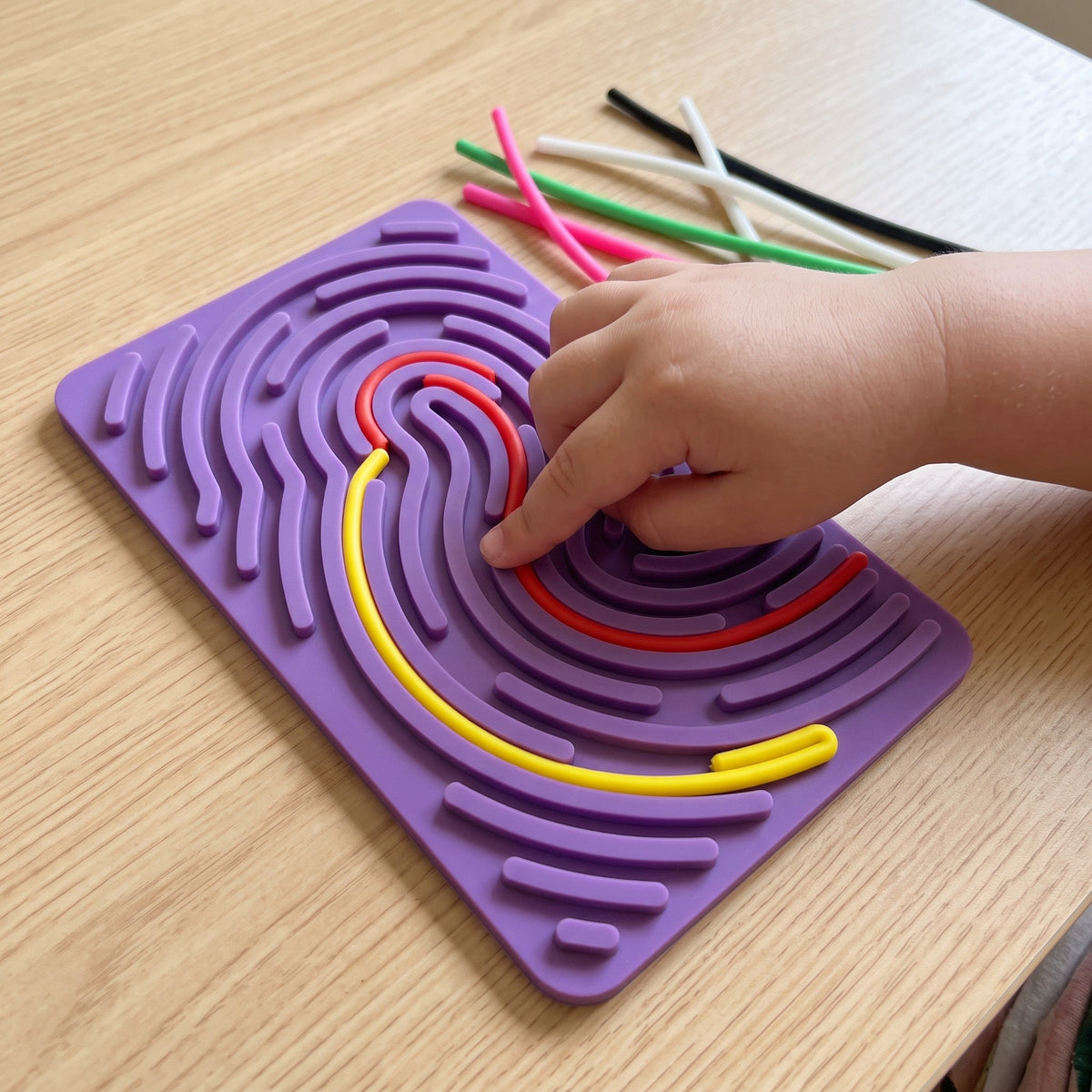 A child's hand presses a red string into the grooves of the purple board, next to a yellow string already in place