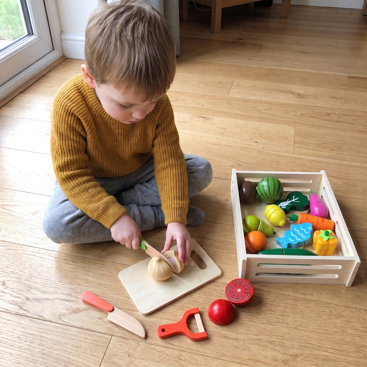 A toddler on the floor cuts a wooden onion on a cutting board, beside the crate of play food, a second knife and a peeler