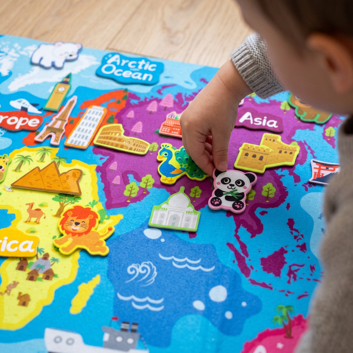 A child's hand presses a felt panda onto Asia, near felt pieces of the Great Wall, the Taj Mahal and a peacock