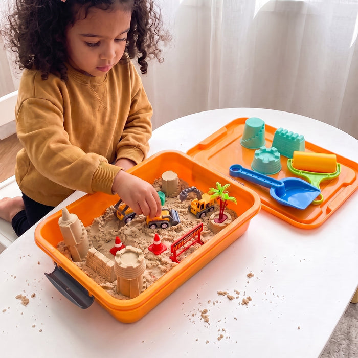 A child plays with toy trucks in the sand bin on a table, with molds, a roller and a shovel resting on the open lid beside it