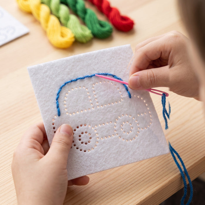 A child's hands pull a pink plastic needle and blue thread through the holes of the car card