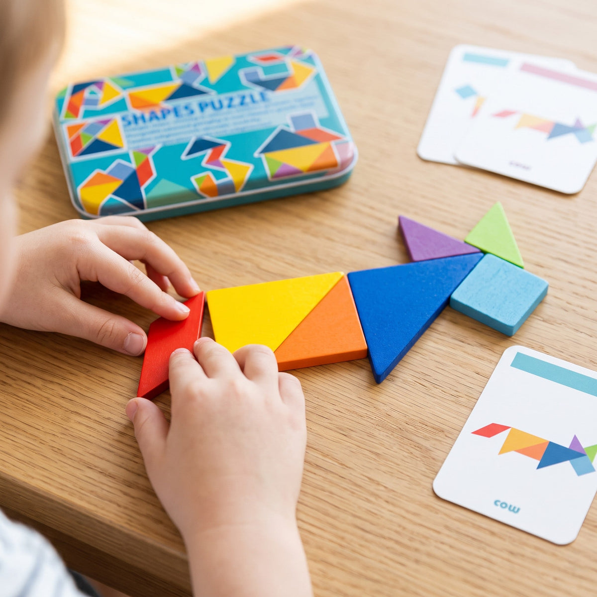 A child's hands build a cow shape from the wooden pieces, following the cow card on the table, with the tin behind