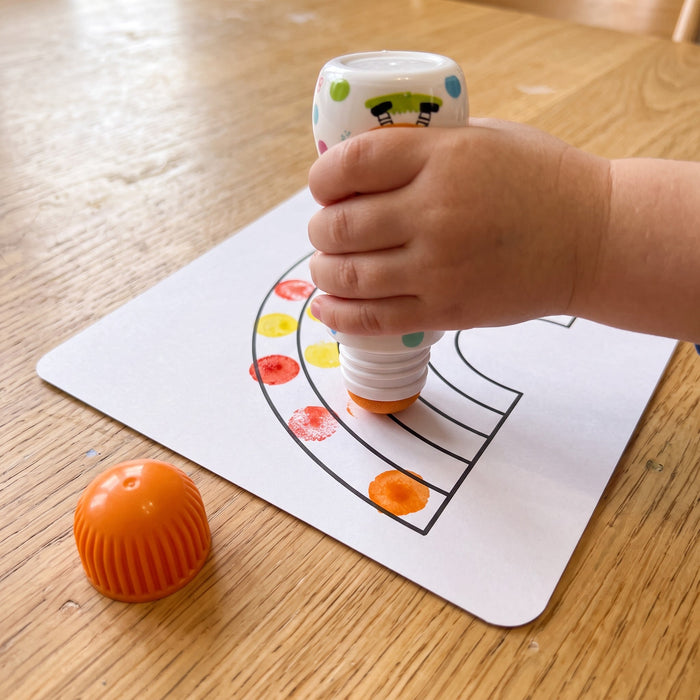A toddler's hand presses an orange dot marker onto a rainbow outline, leaving round dots, with the ribbed cap beside it